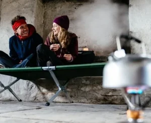 Two campers sitting on a camping cot inside a shelter with a portable stove