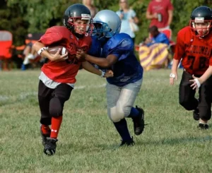 Youth football players in red and blue uniforms during a game, with one player attempting to tackle the ball carrier.