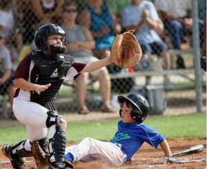 Youth baseball game with a catcher in full gear reaching for the ball as a young player slides into home plate.