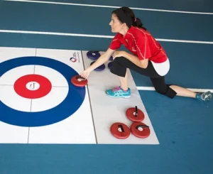 A woman playing floor curling indoors, carefully sliding a red curling stone toward the target on a mat.