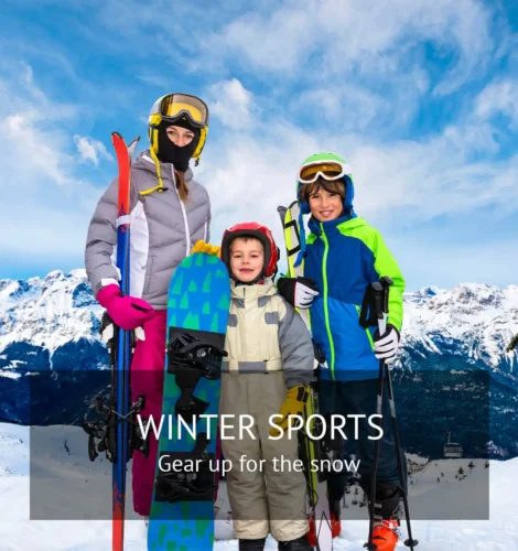 Family of three dressed in colorful winter sports gear, holding skis, a snowboard, and ski poles, standing in front of a snowy mountain range under clear skies.