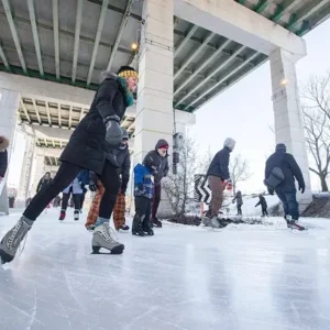 Group of people ice skating under a bridge on a clear winter day, with snow-covered trees in the background.