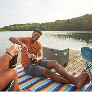 Couple relaxing by a lake with drinks and a cooler on a colorful blanket on a dock.