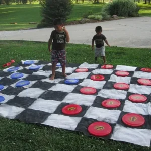 Two children playing a giant outdoor checkers game using colorful discs on a large checkered mat.