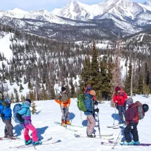 Group of backcountry skiers pausing on a snowy mountain slope, surrounded by evergreen trees and dramatic peaks under a clear sky.