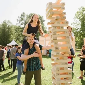 A young woman sitting on a man’s shoulders while playing a giant Jenga game outdoors during a sunny community event.