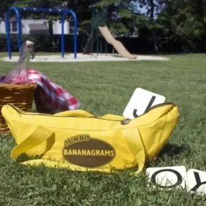 A jumbo Bananagrams yellow bag with oversized letter tiles spilling out, set on a grassy lawn near a picnic area and playground.