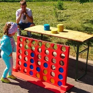 Young girl playing with a giant Connect Four game outdoors, with colorful disks and a table in the background, as an adult takes photos.