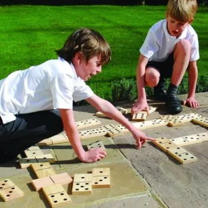 Two children playing with a giant wooden dominoes set on a sunny patio, surrounded by green grass.