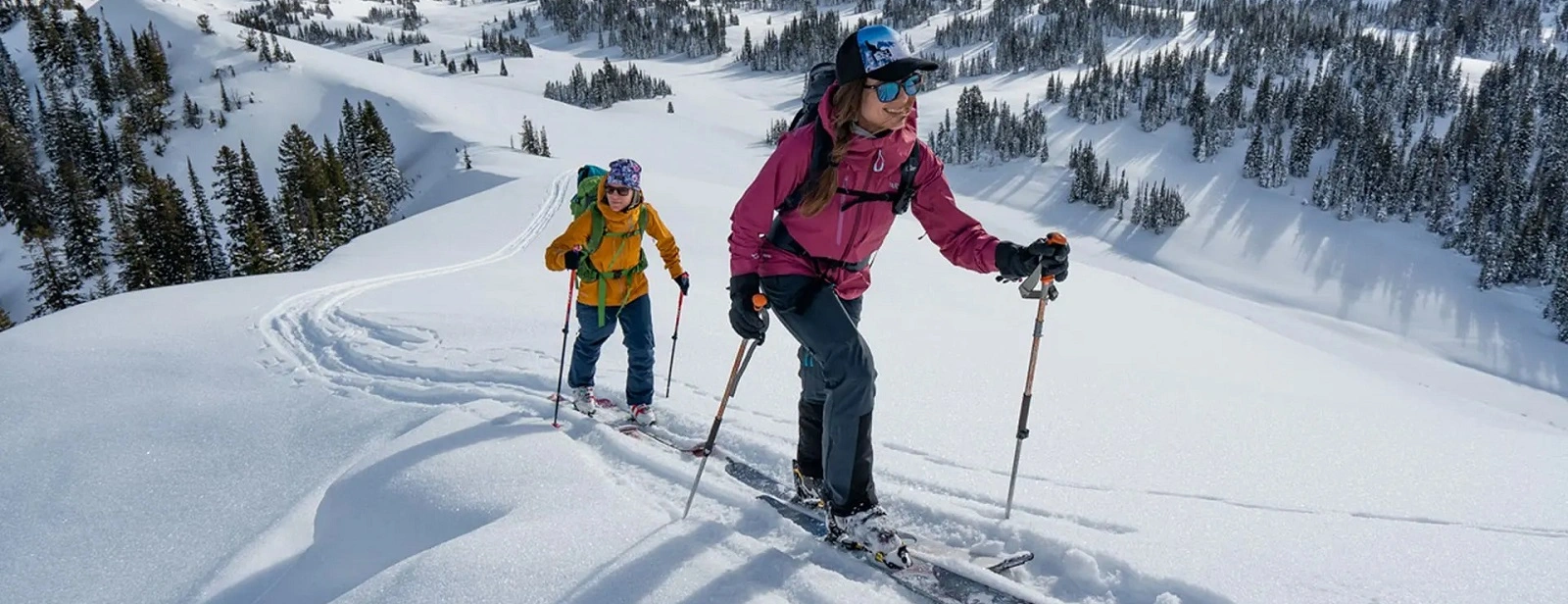 Two women enjoying backcountry ski rentals, skiing uphill in a pristine snowy mountain landscape, wearing bright outdoor gear and carrying backpacks, surrounded by evergreen trees under a clear sky.