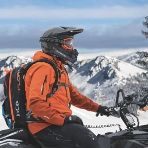 Rider wearing an orange jacket and a snowmobile helmet with a scenic mountain backdrop