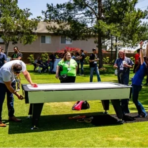 A group of people enjoying an air hockey game outdoors at a sunny event with trees and buildings in the background.