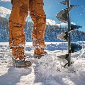 A person standing next to an ice auger while wearing insulated winter boots, surrounded by snowy landscapes.