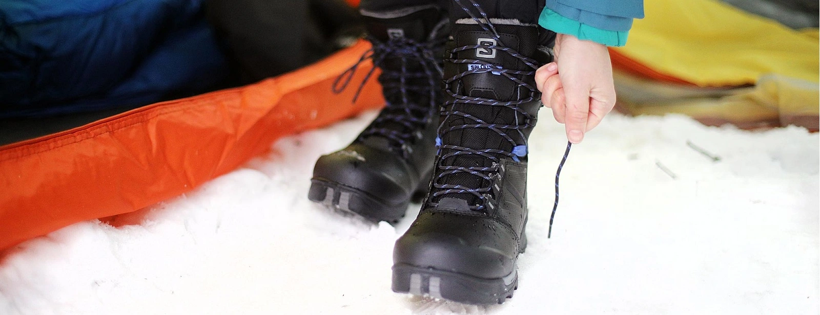 Person tying laces on black Salomon winter boots in the snow near an orange tent. Winter boots for rent in Minnesota