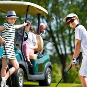 A young boy wearing sunglasses and a white golf outfit prepares to take a shot while two other kids wait by a golf cart.