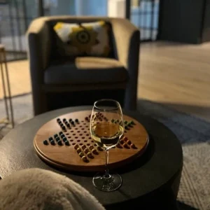 A wooden Chinese Checkers board with a glass of wine resting nearby in a cozy lounge setting.