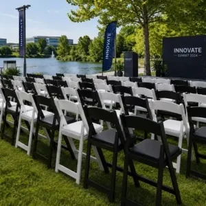 Rows of black and white folding chairs on green grass face a stage with a dark blue backdrop reading 'INNOVATE SUMMIT 2024' beside a lake.